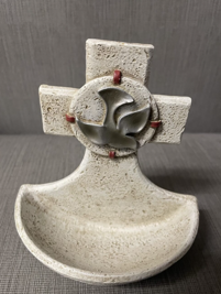 Stone baptismal font with a carved cross and a dove symbol at the center, photographed against a gray background.