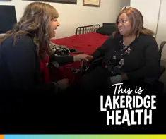 A smiling health-care worker sits beside a woman in a motorized wheelchair in a bedroom, holding her hand in a warm, supportive interaction. The woman sits near a neatly made bed with a red blanket. Text on the image reads “This is Lakeridge Health.”