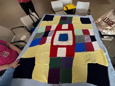 Several people stand around a table holding up a large, handmade knitted blanket made of colourful square panels in yellow, blue, red, green, purple, and white, bordered in light blue.