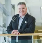 Male doctor smiling indoors, wearing a jacket with a stethoscope around his neck.
