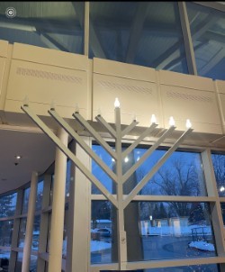Large white menorah with lit candles inside a modern building, with windows and evening light visible outside.