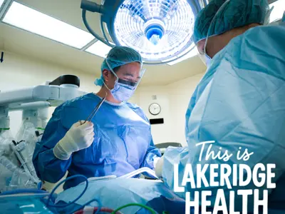 Two surgeons in full protective gear, including masks, gowns, and face shields, perform a procedure under a bright surgical light in an operating room. Medical equipment and monitors surround them. The text ‘This is Lakeridge Health’ appears in the bottom right corner.