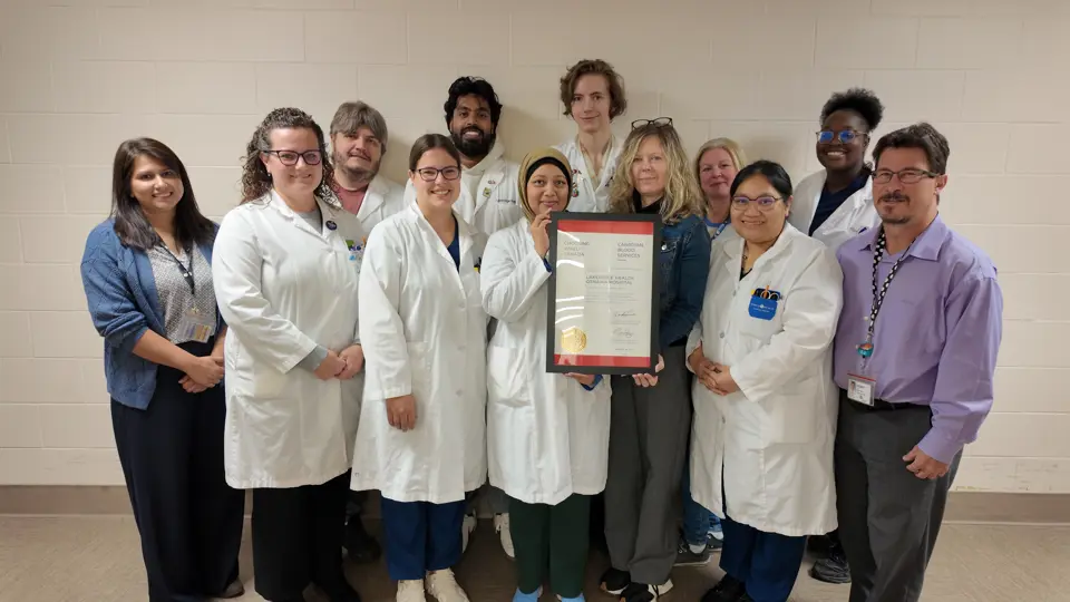 A group of twelve Lakeridge Health team members stand together smiling in a hallway. Several wear white lab coats, and two hold a framed certificate that reads “Choosing Wisely Canada – Canadian Blood Services – Lakeridge Health Oshawa Hospital.”