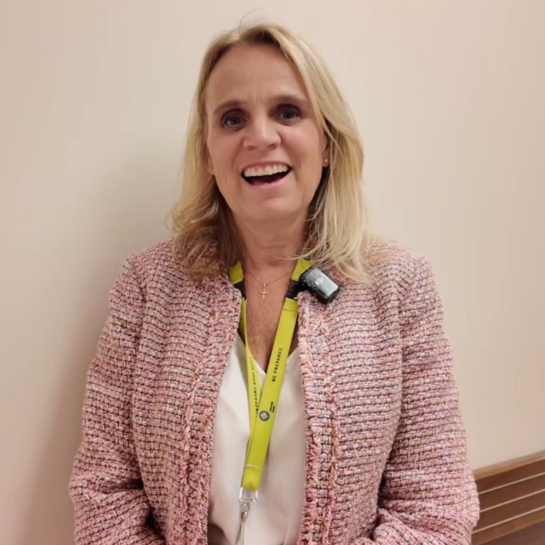 A woman with long blonde hair stands against a neutral wall, smiling broadly. She is wearing a pink textured jacket and a yellow lanyard with an ID badge.