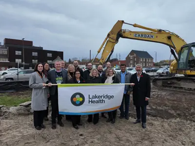 A group of people from Lakeridge Health stand together outdoors at a construction site, smiling and holding a large Lakeridge Health banner.