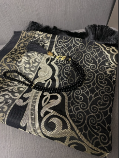 Black and gold patterned prayer mat with tassels, folded on a gray surface, with black prayer beads placed on top.