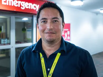 A man stands outside a hospital emergency department entrance, wearing a navy blue shirt and a yellow lanyard. He is smiling slightly, with bright overhead lighting in the background.