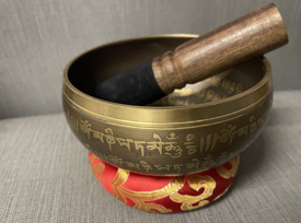 Brass Tibetan singing bowl engraved with script, resting on a red and gold cushion, with a wooden mallet inside.