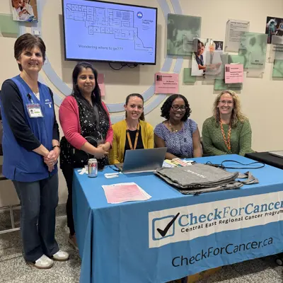 Photo of five women standing and sitting behind a table with a blue tablecloth that says "CheckforCancer.ca"