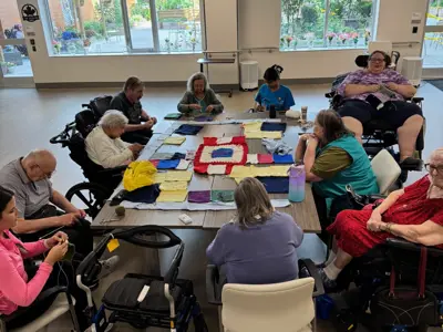 A group of older adults sit and knit around a large table in a bright community space, working together on a colourful patchwork blanket. Walkers and wheelchairs are visible, and large windows look out onto a garden.