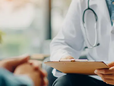 A physician wearing a white lab coat and a stethoscope around their neck holding a clipboard.