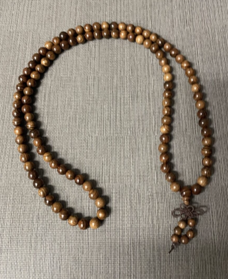 Strand of brown wooden Buddhist prayer beads arranged in a loop on a gray fabric surface, with a small knotted tassel at one end.