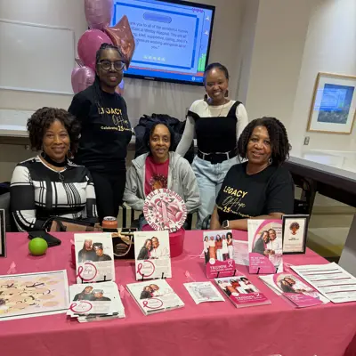 Photo of five women looking at the camera smiling while behind a table with a pink tablecloth