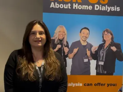 A woman with long brown hair stands in front of a banner that reads “Ask Us About Home Dialysis” with photos of three smiling staff members behind her. She is wearing a black top and a staff lanyard.