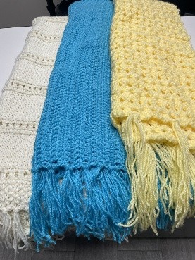 Three knitted prayer shawls in white, blue, and yellow, folded and laid side by side with fringed ends.