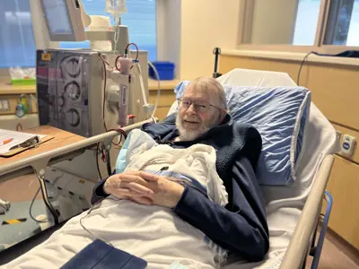 A patient lies in a hospital bed, resting comfortably with hands folded, wearing glasses and covered by a blanket. Medical equipment and monitors are positioned beside the bed, with a window and clinical room furnishings visible in the background.