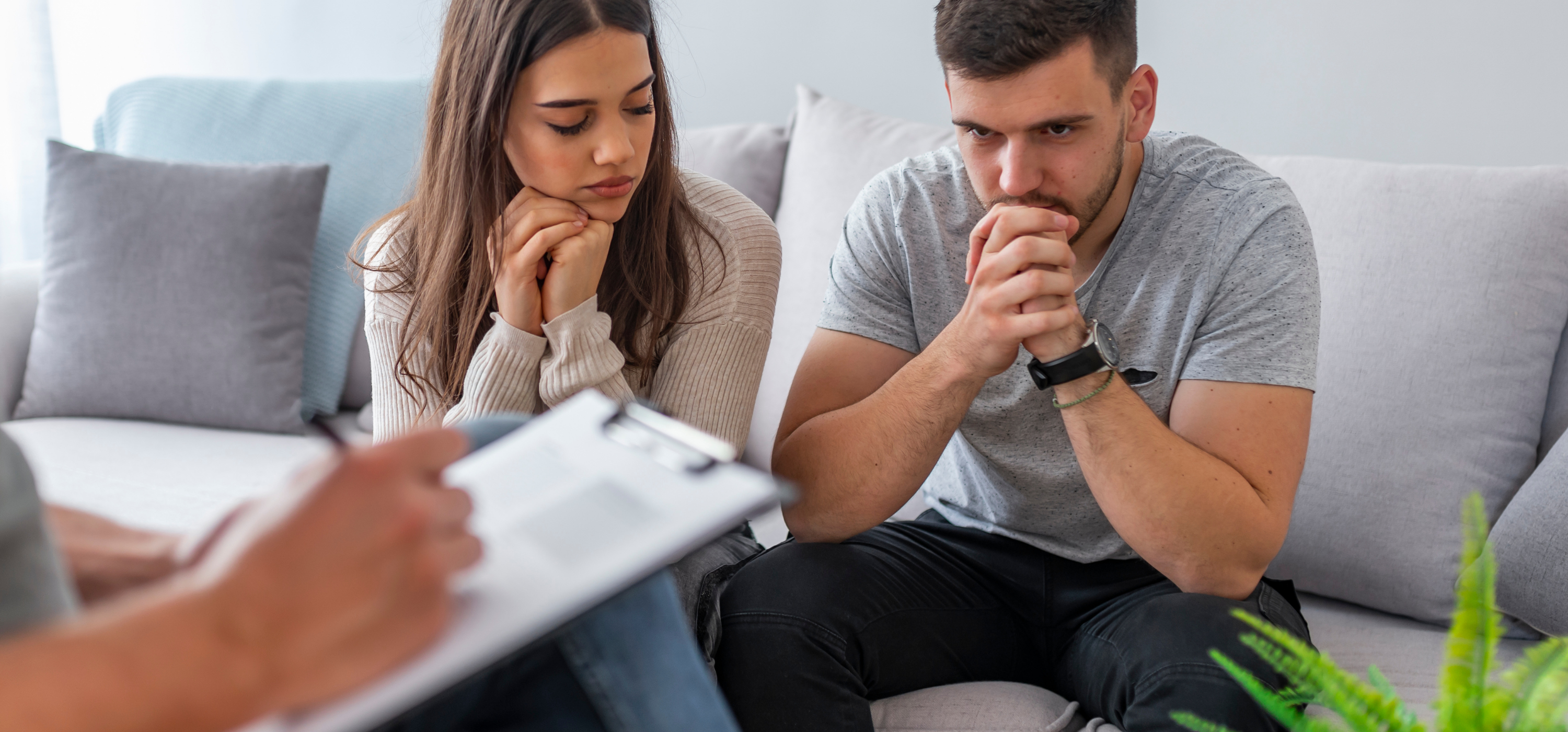 A couple seated on a couch listening. There is a clipboard being held in front of them.