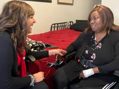 A healthcare professional sits beside a patient in a wheelchair in a bedroom, holding the patient’s hand and speaking with her in a supportive, friendly manner.