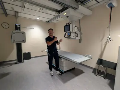 A smiling healthcare professional in dark scrubs stands in a medical imaging room, giving a thumbs-up beside an X-ray machine and examination table. The room has beige walls, overhead equipment, and a clock on the wall.