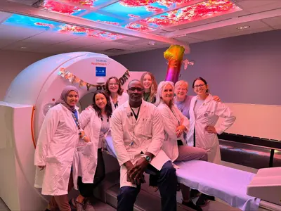 A group of eight medical staff members in white lab coats stand and smile together in front of a large Siemens Healthineers PET CT scanner. A “Happy Birthday” banner is draped across the machine, and the ceiling above glows with colorful panels showing pink blossoms against a blue sky. The group appears cheerful, celebrating in a warmly lit hospital imaging room.